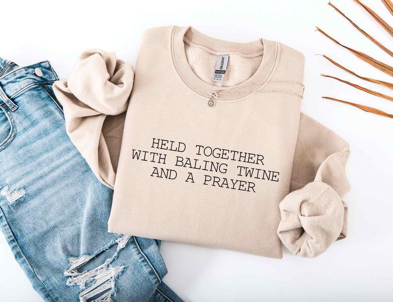 May include: A beige sweatshirt with the text "HELD TOGETHER WITH BALING TWINE AND A PRAYER" in black. The sweatshirt is folded and paired with light blue ripped jeans and a silver necklace.