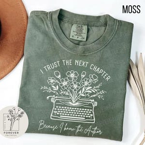 May include: A moss green t-shirt with the text "I Trust The Next Chapter Because I Know The Author" in white. The design features a typewriter with flowers. The shirt is from Forever Faithful Tees.
