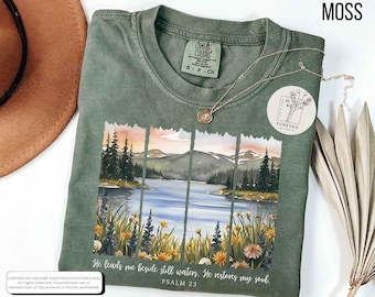 May include: Moss green t-shirt with a watercolor landscape design featuring a lake, mountains, and flowers. The text "He leads me beside still waters. He restores my soul. Psalm 23" is printed below the image. A gold necklace is draped on the shirt.