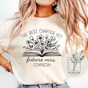 May include: Cream-colored t-shirt with a black floral and book design. The text reads "THE BEST CHAPTER YET future mrs. -JOHNSON-". A small logo with flowers and the words "FOREVER FAITHFUL" is also visible.