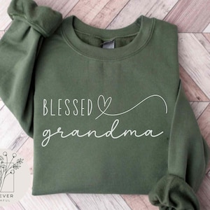 May include: An olive green sweatshirt with the words "Blessed grandma" in white script. A white heart and curved line design is above the word "grandma". The sweatshirt has long sleeves and a crew neck.