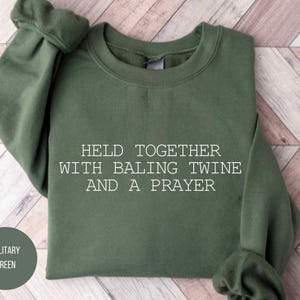 May include: Military green sweatshirt with the text "HELD TOGETHER WITH BALING TWINE AND A PRAYER." The sweatshirt is folded on a light wood-grain surface. A circular label in the lower left corner reads "MILITARY GREEN."