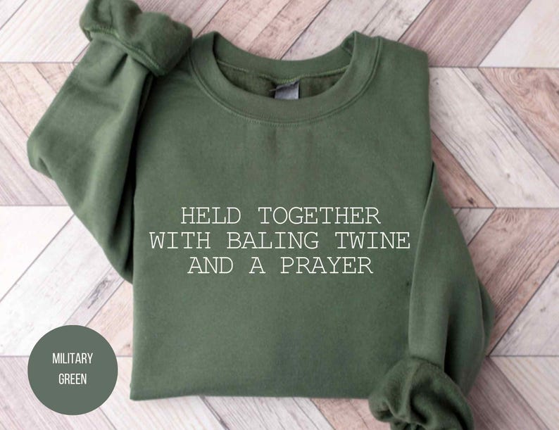 May include: Military green sweatshirt with the text "HELD TOGETHER WITH BALING TWINE AND A PRAYER." The sweatshirt is folded on a light wood-grain surface. A circular label in the lower left corner reads "MILITARY GREEN."