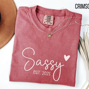 May include: A crimson red t-shirt with the text "Sassy est 2025" in white script with a small pink heart.