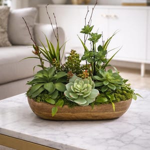 May include: A wooden oval planter filled with assorted faux succulents and greenery. The arrangement includes various shades of green plants, moss, and small branches. The planter sits on a marble-topped coffee table with a gold base.