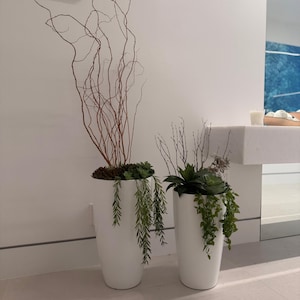 May include: Two tall, white, cylindrical planters containing artificial greenery. The taller planter features long, thin, brown, twisted branches and trailing green foliage. The shorter planter holds smaller twigs and a cluster of green succulents.