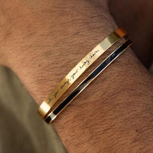 Peut inclure: Deux bracelets joncs en métal doré. L'un des bracelets présente une surface lisse et polie, tandis que l'autre est gravé du texte en cursive noire, "oh you always, your lucky charm". Les deux bracelets ont un design fin et ouvert.