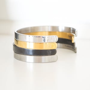 May include: Four stacked metal bracelets in silver, gold, black, and silver. Each bracelet has a small engraved design.