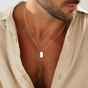May include: A silver-toned necklace featuring a delicate chain and a small, smooth, rectangular pendant. The pendant hangs centered on the chain, which rests against the neck. The metal has a polished, reflective finish that catches the light.