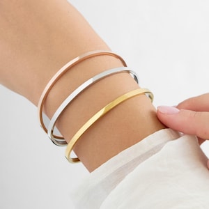 May include: Three thin metal bracelets stacked on a wrist. The bracelets are gold, silver, and rose gold.