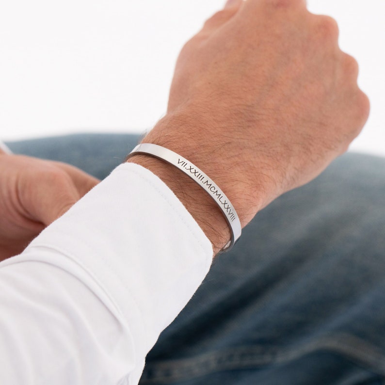 May include: A silver cuff bracelet with the Roman numeral date VII.XXIII.MCMLXXXVIII engraved on it.