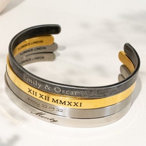 May include: A layered cuff bracelet with a black, gold, and silver finish. The bracelet is engraved with the text "Emily & Oscar", "XII XII MMXXI", "Poppy 22.09.22", and "Monty".