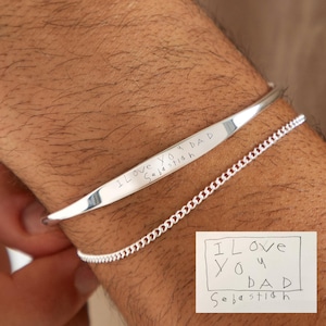 Peut inclure: Un bracelet en argent gravé avec l'inscription "I LOVE YOU DAD Sebastian". Un second bracelet chaîne en argent plus fin est également visible. Une note manuscrite avec le même message est dans le coin inférieur droit.