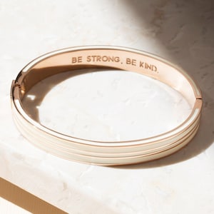 May include: A rose gold bangle bracelet with a white enamel band and the inscription "BE STRONG. BE KIND."