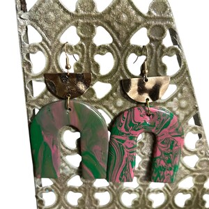 May include: A pair of abstract earrings with a gold-tone half-circle top and a marbled arch design. One earring features green and pink swirls, while the other has a pink and green pattern. The earrings are displayed on a decorative metal stand.