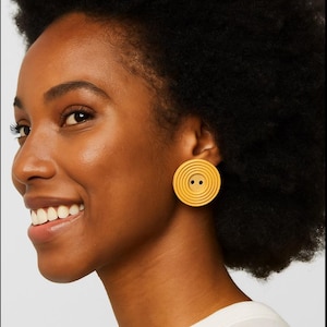 May include: A close-up of a person wearing a pair of yellow circular earrings. The earrings have a spiral design with two small black dots in the center. The earrings are a bright, solid yellow color.