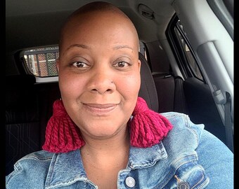 May include: A woman with a shaved head wearing a denim jacket and large pink tassel earrings.