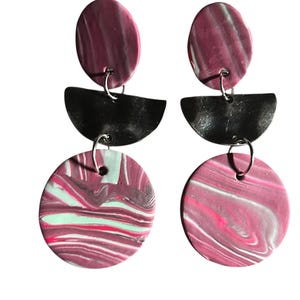 May include: A pair of handmade earrings with a layered design. The top oval is a marbled burgundy, the middle is a black half-circle, and the bottom is a marbled circle with burgundy, pink, and mint green. The earrings hang from silver hooks.