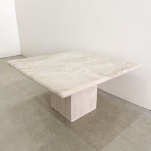May include: A square travertine coffee table with a light beige color and a simple, modern design. The table has a square top and a single square base.