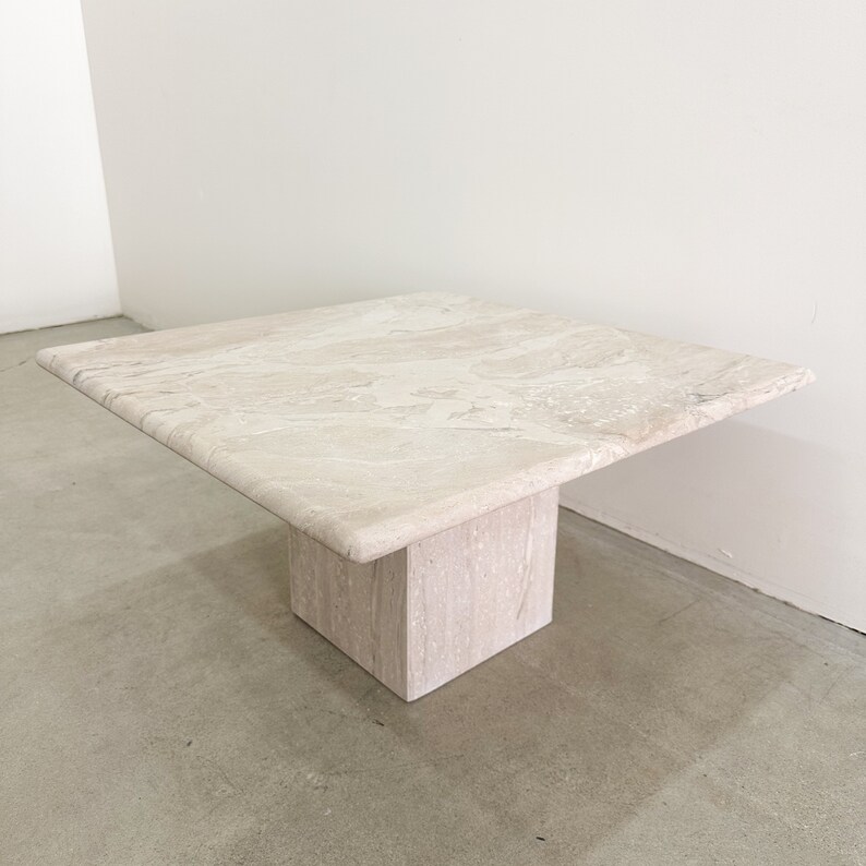 May include: A square travertine coffee table with a light beige color and a simple, modern design. The table has a square top and a single square base.