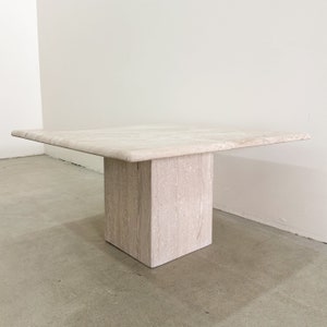 May include: A square, light beige travertine coffee table with a thick, square base. The table has a smooth, polished surface.