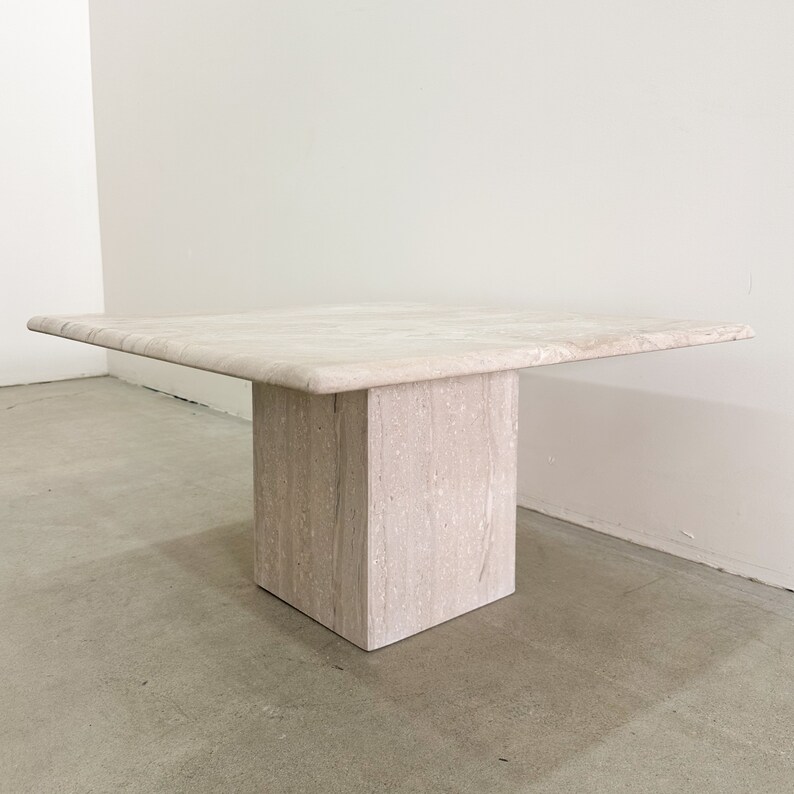 May include: A square, light beige travertine coffee table with a thick, square base. The table has a smooth, polished surface.