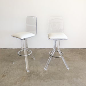 May include: Two clear acrylic bar stools with white padded seats and chrome bases.