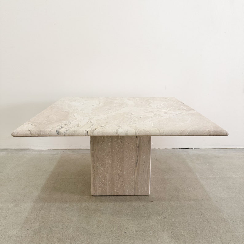 May include: A square travertine coffee table with a light beige color and a natural stone finish. The table has a single square base.