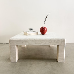 May include: A square, light-colored stone coffee table with a minimalist design. A red vase with a branch, a small glass cup, and a stack of books are on the table. The table has four thick, rectangular legs.