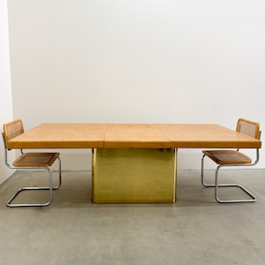 May include: A rectangular dining table with a brown leather top and a gold metal base. Two chrome chairs with woven cane seats are placed on either side of the table.
