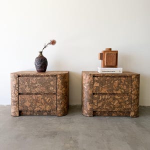 May include: Two matching nightstands with a cork veneer finish. Each nightstand has two drawers and rounded edges. One nightstand has a dark brown vase with a dried flower, and the other has a wooden sculpture and a book.