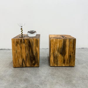 May include: Two wooden cube side tables with a natural wood grain finish. One table has a martini glass with a gold stem and a small glass bowl. The tables are on a concrete floor.