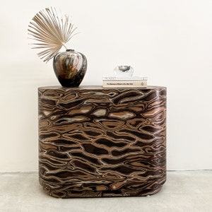 May include: A brown and black patterned side table with a rounded top and a book titled "Picasso" and a clear glass object on top. A dried palm leaf and a brown and white ceramic vase are on the table.
