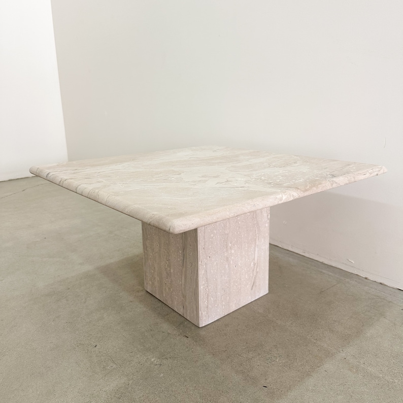 May include: A square travertine coffee table with a light beige color and a natural stone finish. The table has a square top and a single square base.