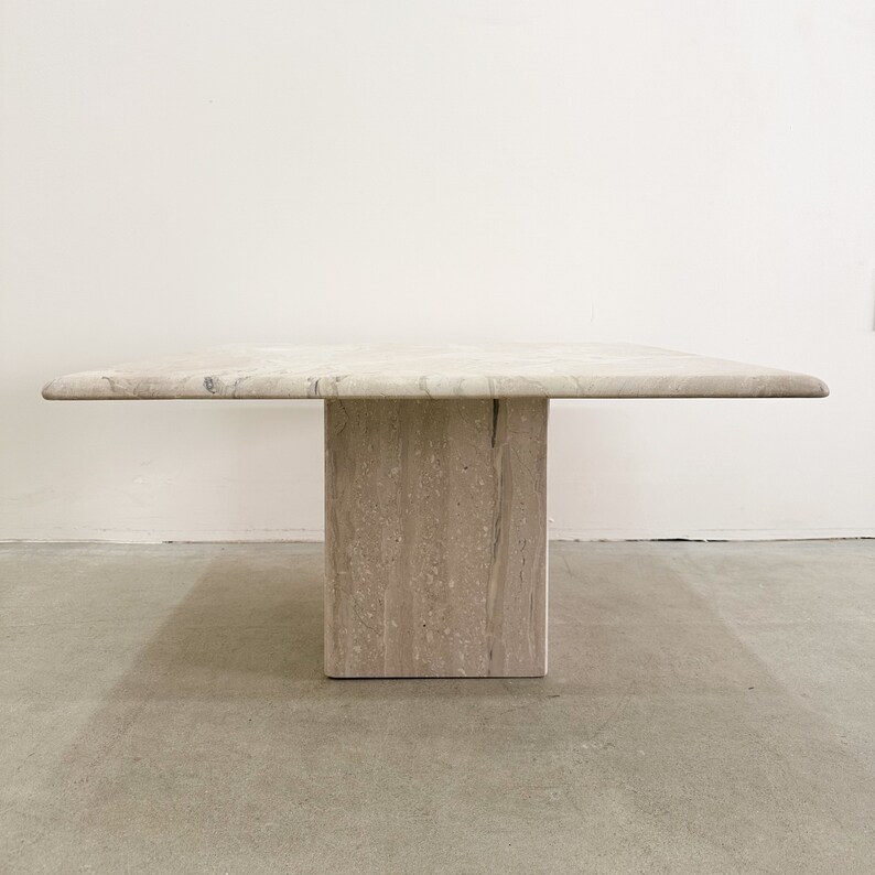 May include: A square travertine coffee table with a light beige color and a single, thick, square leg.