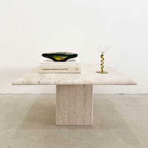 May include: A white travertine coffee table with a square top and a single pedestal base. A green glass bowl and a book titled "Picasso" are on the table. A clear glass with a gold spiral stem is also on the table.