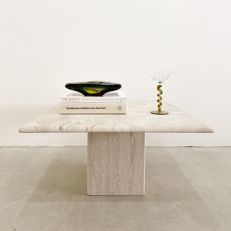 May include: A white travertine coffee table with a square top and a single pedestal base. A green glass bowl and a book titled "Picasso" are on the table. A clear glass with a gold spiral stem is also on the table.