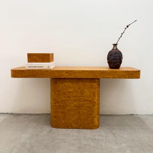 May include: A light brown wooden console table with a rectangular top and a solid, curved base. The table features a decorative wood grain pattern. A small wooden box and two books are on the left side, and a dark brown vase with a branch is on the right.