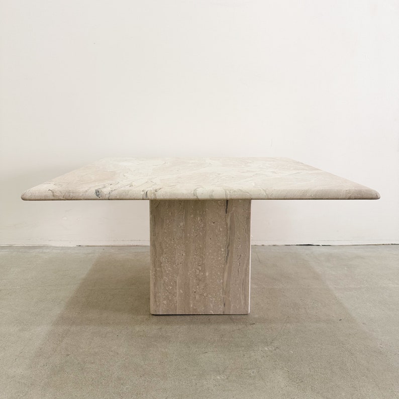 May include: A square travertine coffee table with a light beige color and a natural stone finish. The table has a single square base.