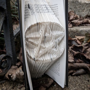 Op de afbeelding: Een boek gevouwen tot een driedimensionaal origami-achtig beeld van een vogel. Het boek is open en de pagina's zijn gevouwen om de vorm van de vogel te creëren. Het boek ligt op een bruin oppervlak.