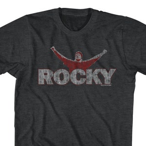 May include: A dark heather grey t-shirt with a distressed red graphic of a man with arms raised and the word "ROCKY" in red letters.