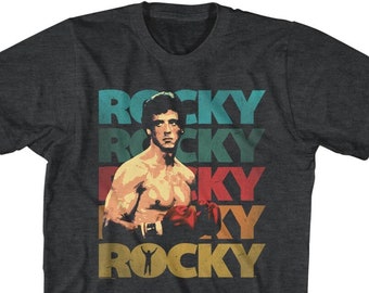 May include: A dark heather grey t-shirt with a vintage-style graphic of Sylvester Stallone as Rocky Balboa. The graphic features the word "ROCKY" repeated in different colors and fonts, with a silhouette of Rocky raising his fists in victory.