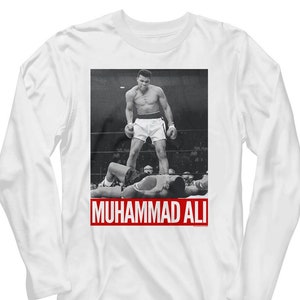 May include: White long-sleeve t-shirt with a black and white photo of Muhammad Ali standing over a fallen opponent. The text "MUHAMMAD ALI" is printed in red at the bottom of the image.