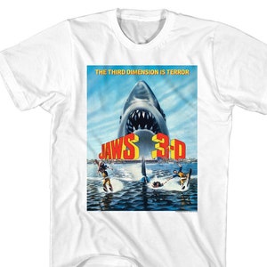 May include: White t-shirt with a Jaws 3-D movie poster print. The poster features a large great white shark with its mouth open, and the text "The Third Dimension is Terror" and "Jaws 3-D" in red and yellow letters.
