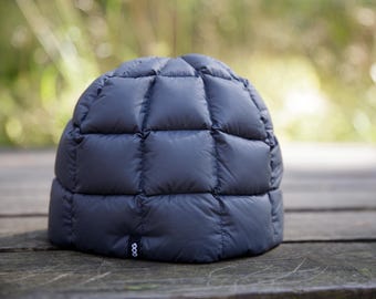 Puede incluir: Un gorro acolchado de color gris oscuro con un patrón de cuadrícula de cuadrados cosidos. El gorro presenta una pequeña etiqueta de tela negra en el borde inferior con el texto blanco GOG impreso. El material parece suave y mullido con un acabado liso.