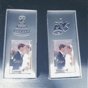 May include: Two silver picture frames with a wedding theme. The top of the left frame has a silver cake design and the top of the right frame has two silver bells. Both frames have a clear photo insert of a bride and groom.