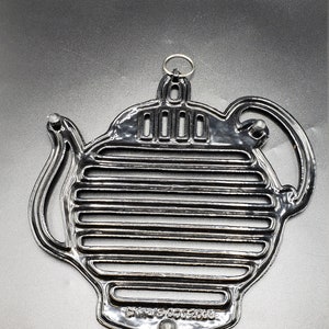 Puede incluir: Un salvamanteles de metal plateado con forma de tetera. El salvamanteles tiene un asa, un pico y una serie de barras horizontales. Un pequeño anillo está unido para colgar. Diseñado para proteger las superficies de la cocina caliente.