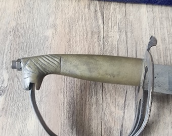 May include: A close-up of the handle of a sword with a brass hilt and a leather strap. The sword is on a wooden surface.