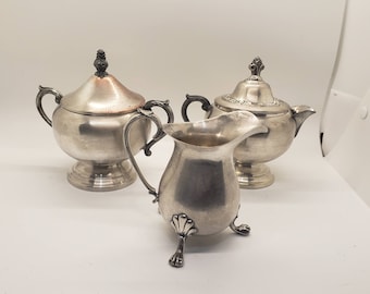 Puede incluir: Juego de té de plata de tres piezas. El juego incluye un azucarero con tapa, una jarra para crema y una tetera con tapa. Las piezas tienen asas ornamentadas y detalles decorativos. El juego se exhibe sobre una superficie blanca.