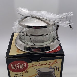 May include: A silver condiment jar set with a lid and a spoon. The set is packaged in a black box with the words "TableCraft" and "Condiment Jar Set" printed on it. The box also includes the text "Stainless steel, Spoon included, Ideal for grated cheese, sugar, relish, horseradish, mustard, mayonnaise".